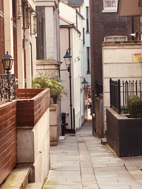A narrow urban alleyway with paved stone flooring situated between residential buildings, featuring a mix of brick and painted facades. On the left, there is a wooden planter box with some greenery and a traditional black metal lamp post mounted on the wall. On the right, a black metal fence with vertical bars encloses a small garden area, alongside another building with a concrete wall and a signage plaque. Further down the alley, a larger vehicle, likely a moving van, is partially visible, indicating a loading or unloading process related to home relocation services. The scene suggests a residential area suitable for house removals and furniture transport, with Brompton Movers possibly involved in managing the logistics of this move through a narrow street environment.