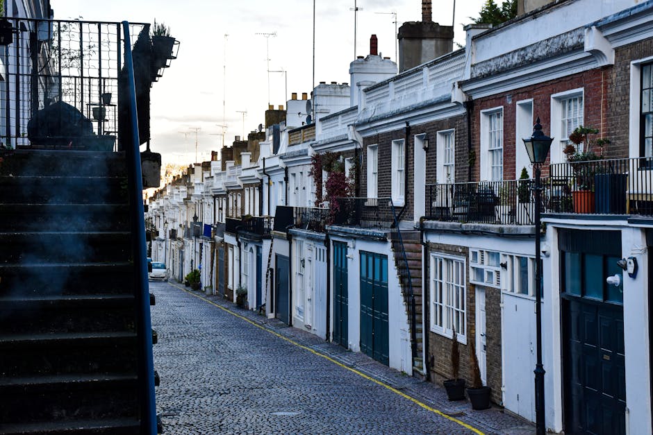 A narrow urban alleyway with paved stone flooring situated between residential buildings, featuring a mix of brick and painted facades. On the left, there is a wooden planter box with some greenery and a traditional black metal lamp post mounted on the wall. On the right, a black metal fence with vertical bars encloses a small garden area, alongside another building with a concrete wall and a signage plaque. Further down the alley, a larger vehicle, likely a moving van, is partially visible, indicating a loading or unloading process related to home relocation services. The scene suggests a residential area suitable for house removals and furniture transport, with Brompton Movers possibly involved in managing the logistics of this move through a narrow street environment.