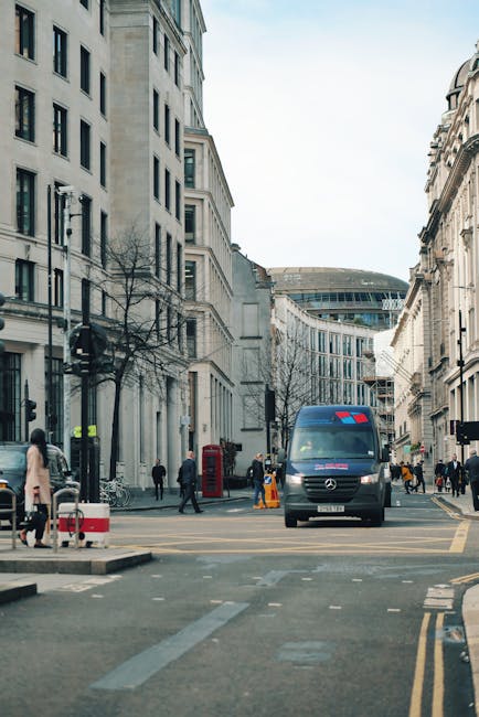 Exterior view of a busy London street on a clear day, showing multi-storey buildings with classic architecture lining both sides. A black Mercedes-Benz van with red markings is parked on the road, part of the moving process associated with home relocation. Several pedestrians are walking on the sidewalk, some passing by and others approaching the van. On the pavement, there are orange traffic cones and barriers indicating ongoing loading or unloading activities. Nearby, a red telephone booth and street furniture are visible, with leafless trees lining the street. The scene suggests the transportation of furniture and packed boxes, contributing to a house removal project. Brompton Movers is involved in the professional packing and furniture transport services, supporting client logistics on busy weekends such as this.
