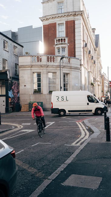 A cyclist wearing a red helmet and pink jacket rides a black bicycle across a busy urban intersection on Old Brompton Road during daylight. In the background, a white delivery van with the initials 'RSL' parked alongside a classic multi-storey building featuring white stone and red brick façade, decorative balcony railing, and tall windows. The street scene includes parked cars, pedestrian crossing lines, and bollards, with natural sunlight illuminating the scene. The setting reflects a typical street environment where house removals or furniture transport logistics might occur, with Brompton Movers providing moving services that involve loading and unloading furniture and boxes within this area.